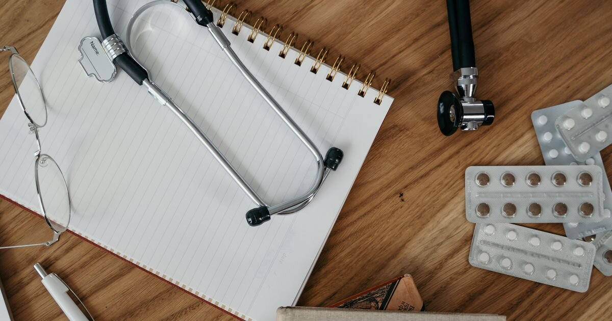 Stethoscope resting on a notebook beside pill blister packs on a wooden desk. Sorting a healthcare stocks list
